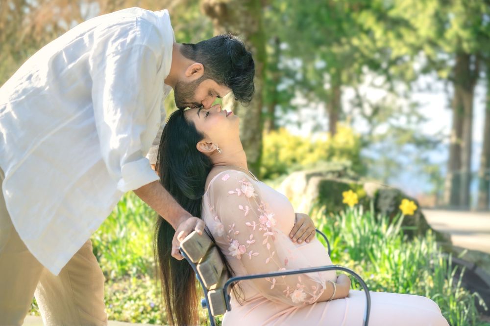 A man tenderly kisses a pregnant woman outdoors in a sunlit park during a maternity photo shoot.