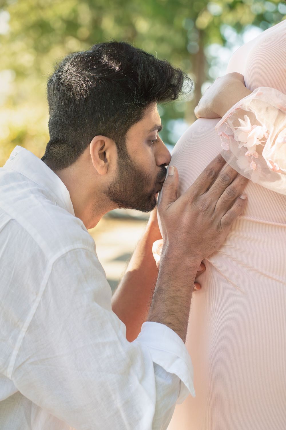 Man kissing pregnant woman's belly outdoors, celebrating pregnancy with a pink bow decoration.
