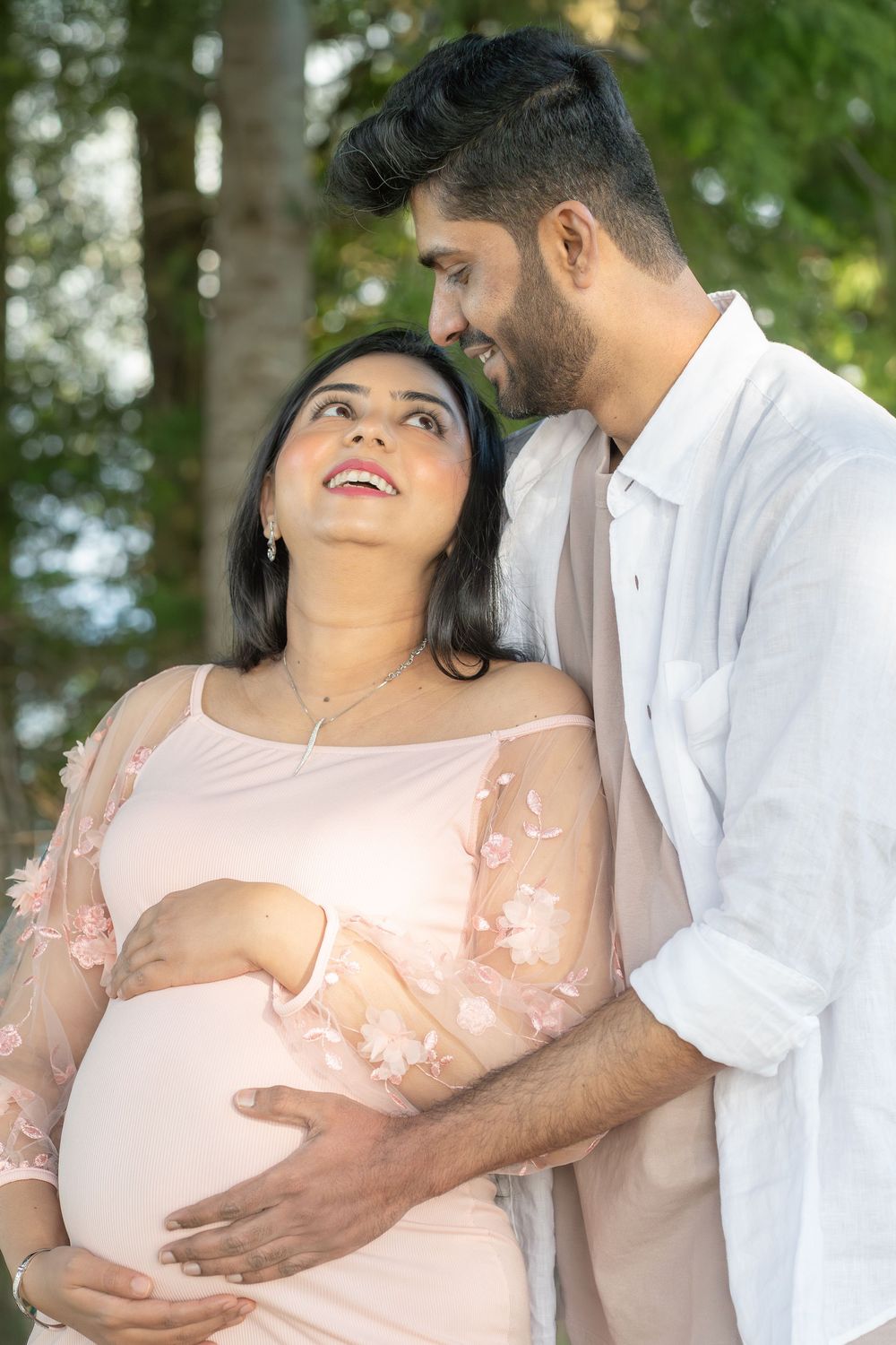 A smiling pregnant woman in a pink dress with her partner gently cradling her baby bump outdoors.