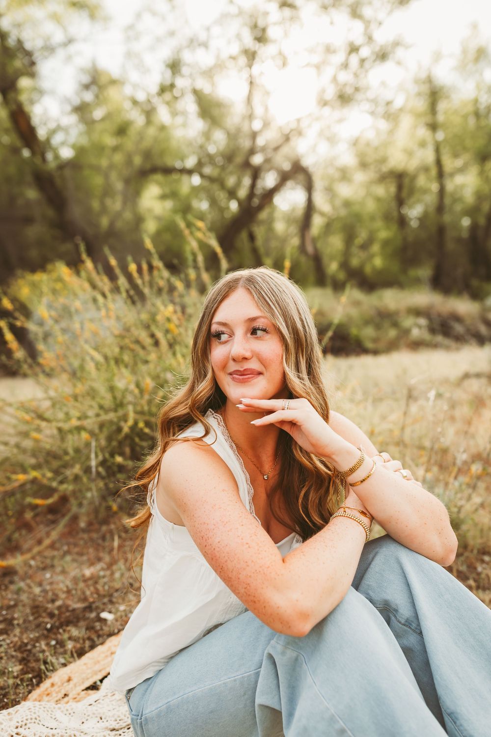Young woman with long blonde hair sits outdoors in jeans and white top, smiling in a sunlit natural setting.