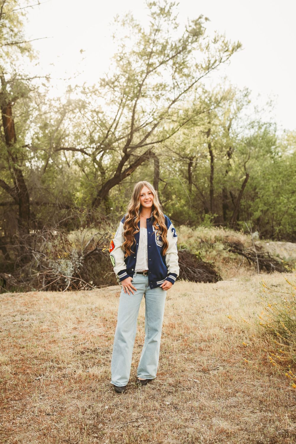 Smiling teen girl in varsity jacket and light jeans stands on dry grass with trees in background.