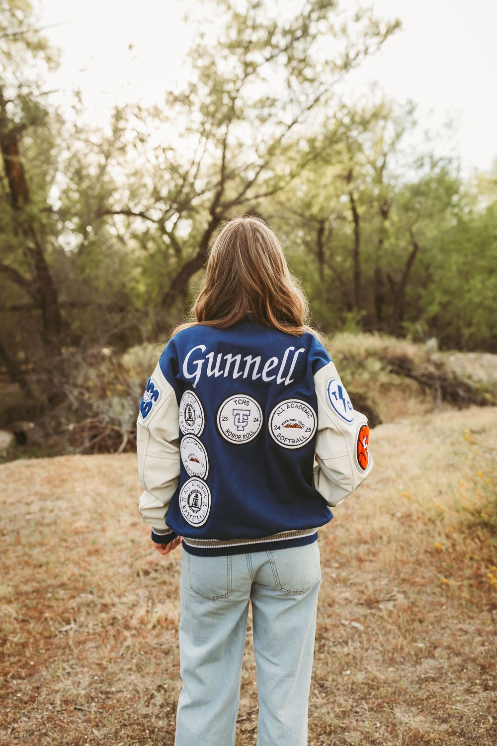 Person wearing a navy Gunnell varsity jacket with patches, standing in a natural outdoor setting.