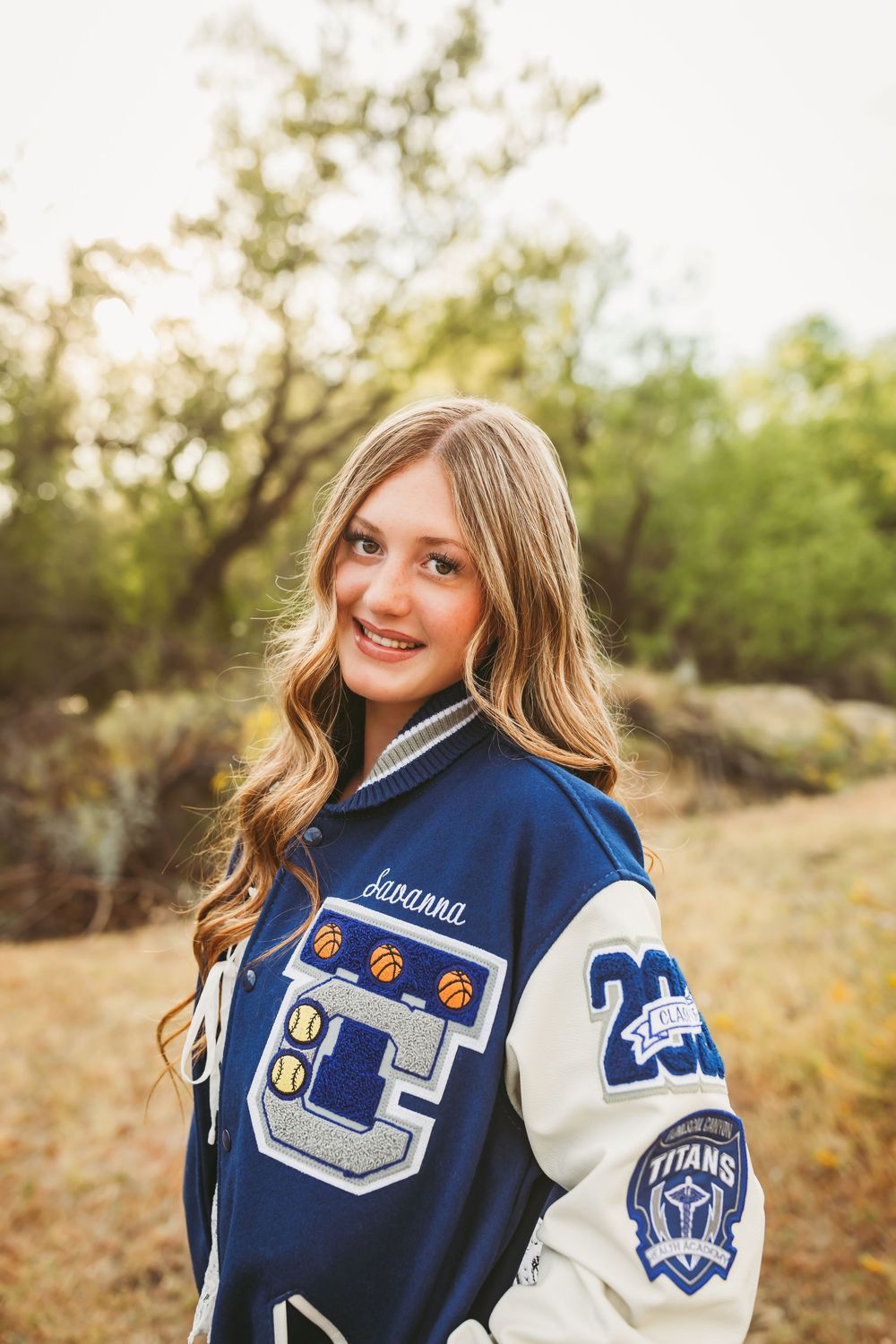 Smiling teen girl wearing a blue and white Titans letterman jacket outdoors with trees in background.