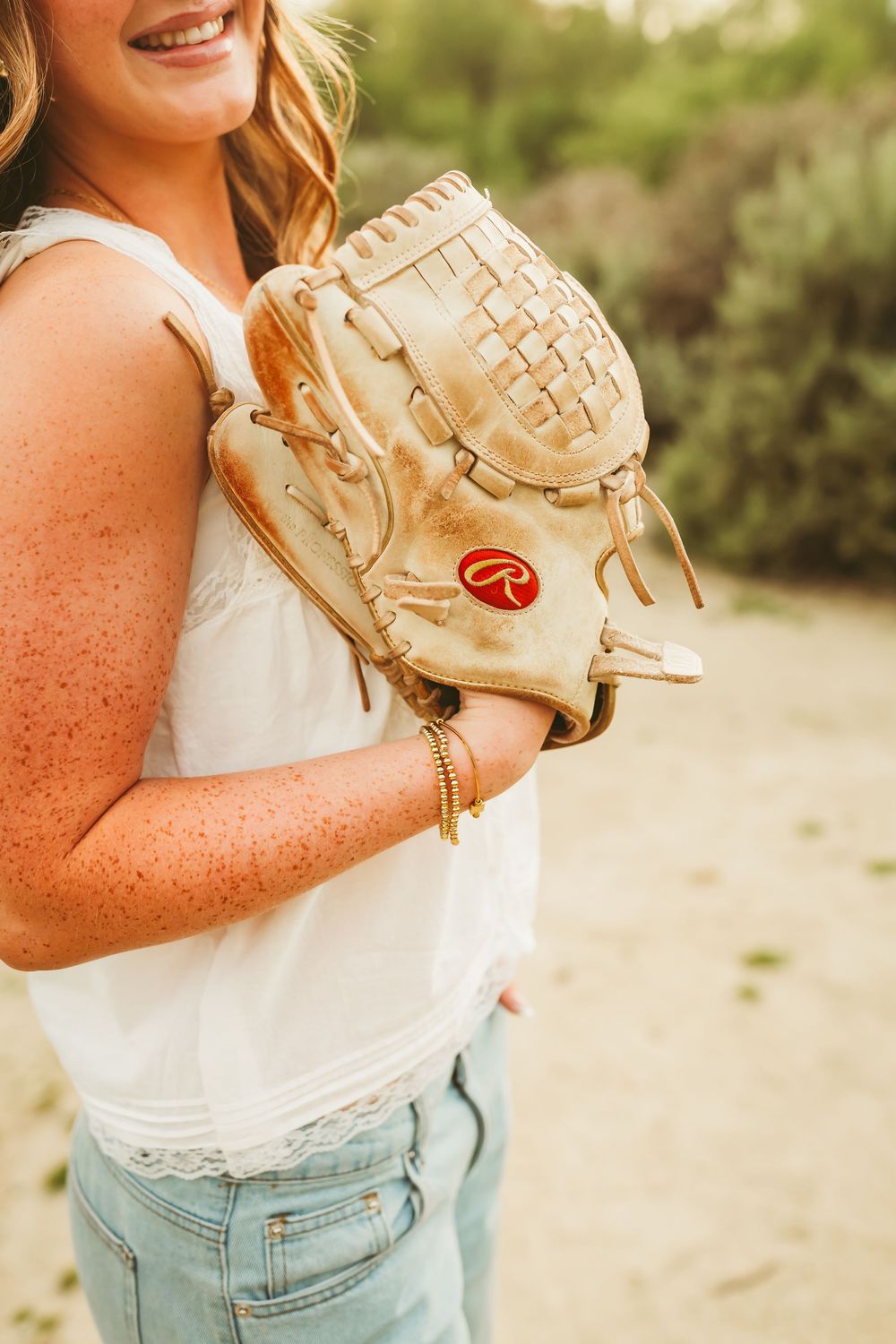 Young woman holding a Rawlings baseball glove while wearing a white top and light jeans outdoors.