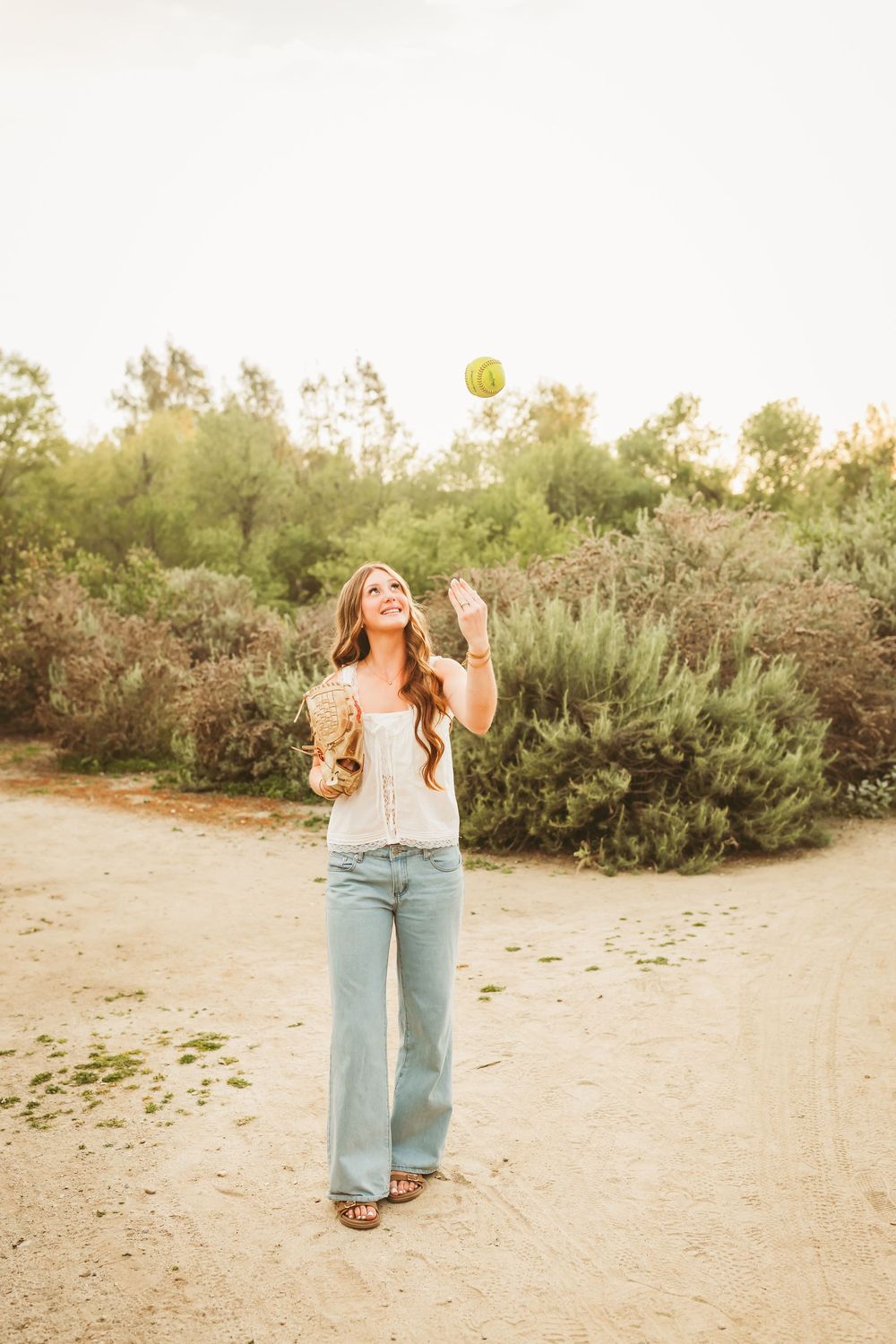Smiling young woman in white top and wide-leg jeans tossing a tennis ball outdoors on a dirt path.