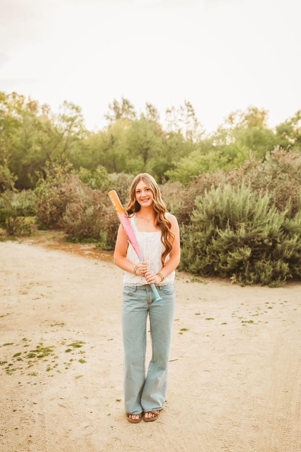Smiling teen girl holding colorful softball bats on a dirt path surrounded by green shrubs.
