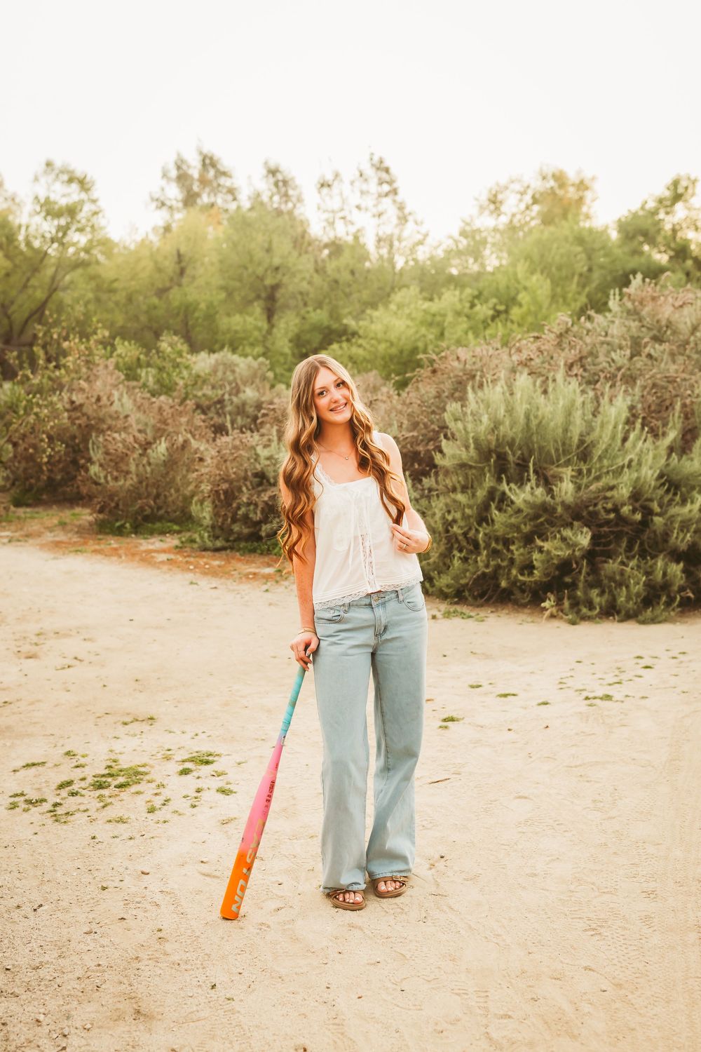 Teen girl with wavy hair holding a colorful softball bat on a sandy path surrounded by greenery.