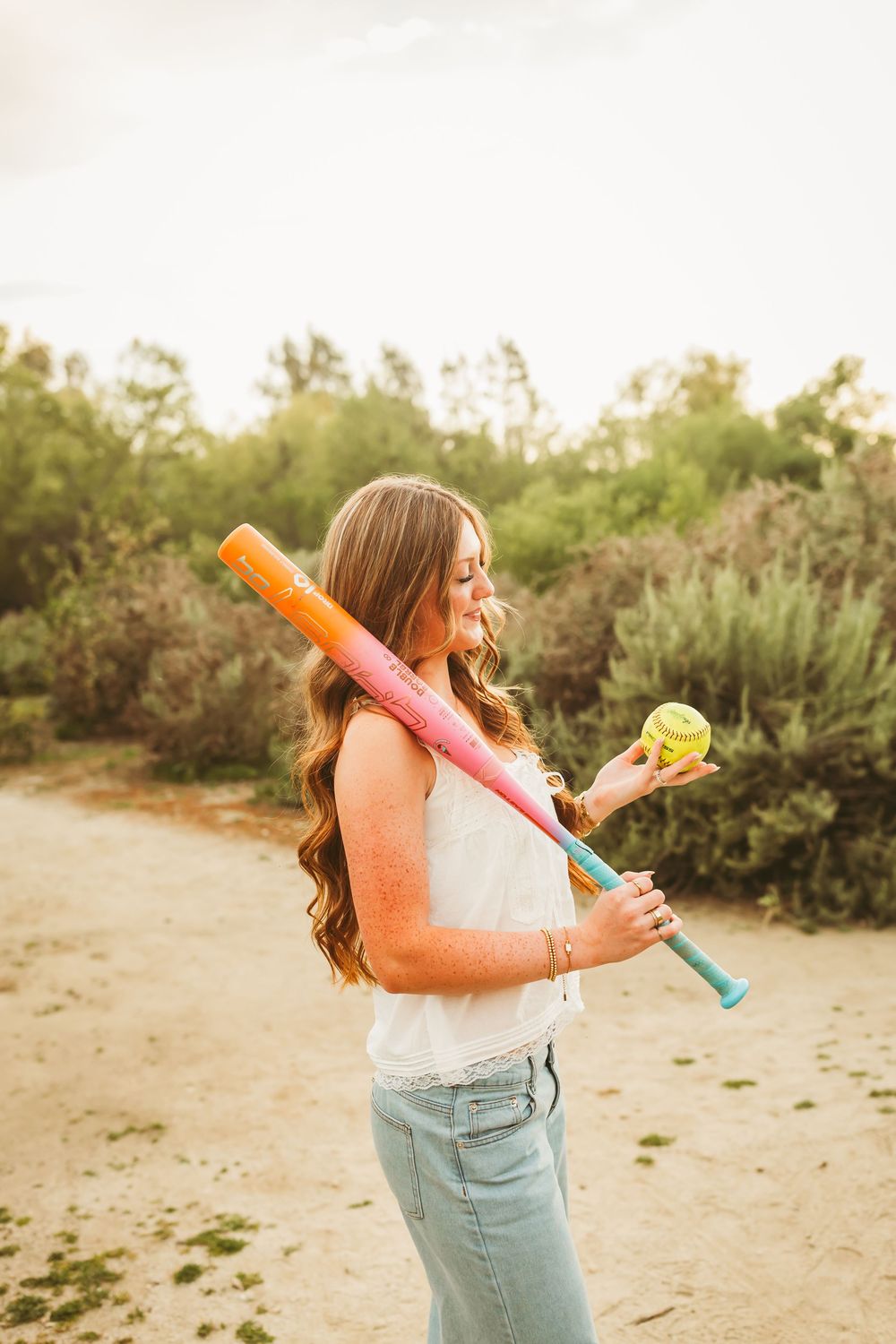 Young woman in white top and jeans holding a colorful bat and softball ball outdoors in nature.