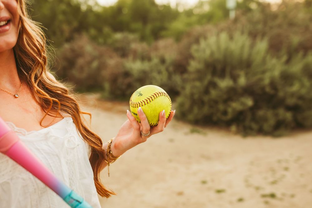 Young woman with long wavy hair holding a yellow softball outdoors near green shrubs on a sandy field.