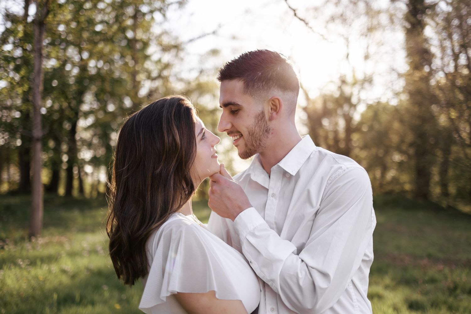 Romantic couple sharing a tender moment outdoors in a sunlit forest, man in white shirt gently touching woman's chin.