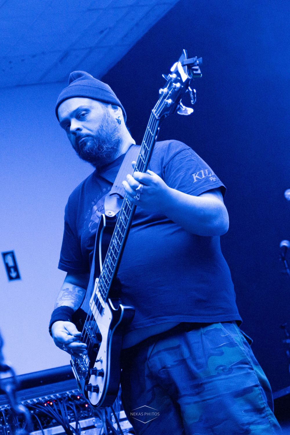 Bearded guitarist in blue beanie plays electric bass guitar on stage under blue lighting at a live music concert.