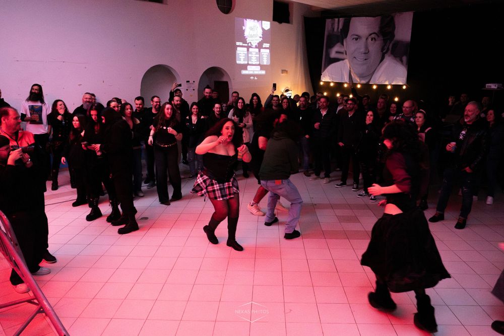 People dancing at a lively indoor event with a crowd watching under pink lights and a projected image on the wall.