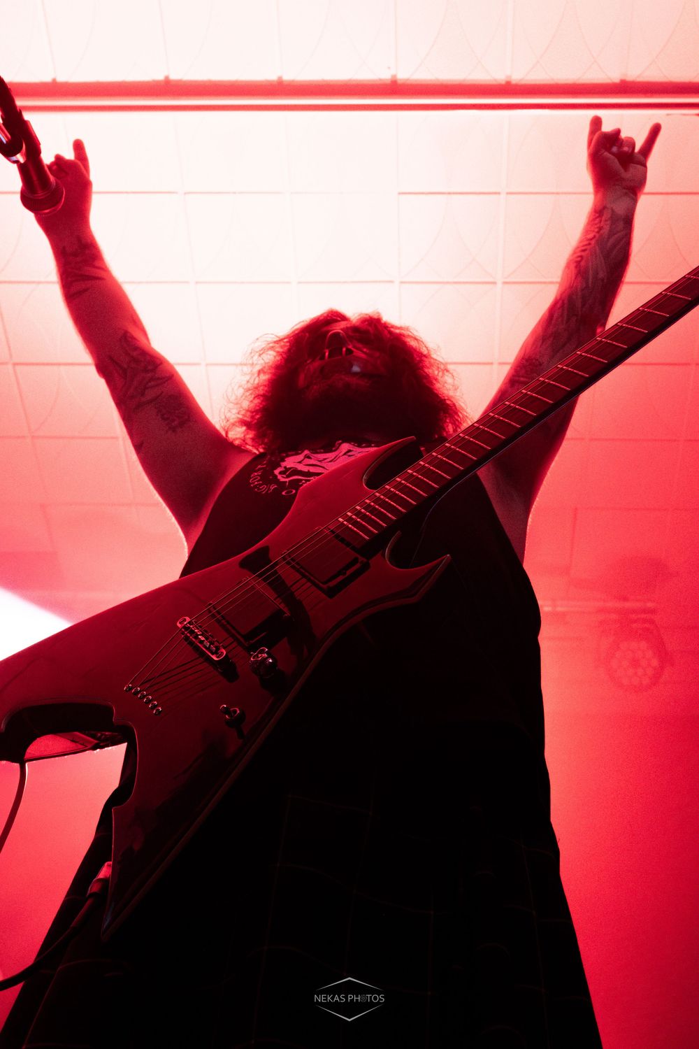 Tattooed metal guitarist raises devil horns under red stage lighting during live concert performance.