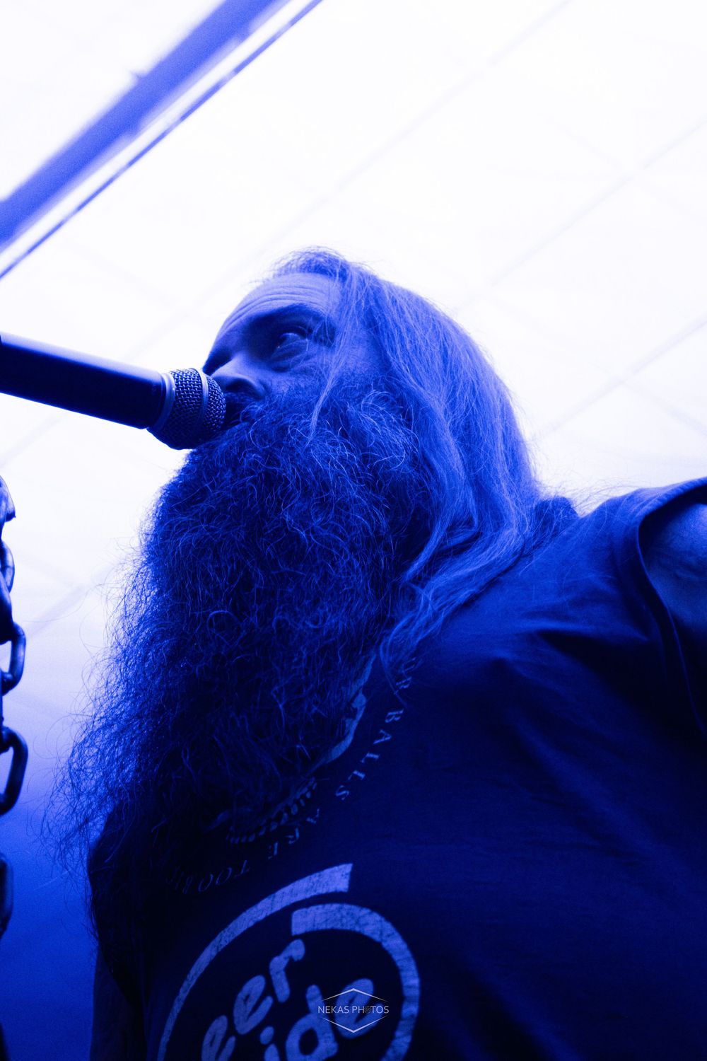 A long-haired, bearded metal vocalist performs on stage under dramatic blue lighting while singing into a microphone.