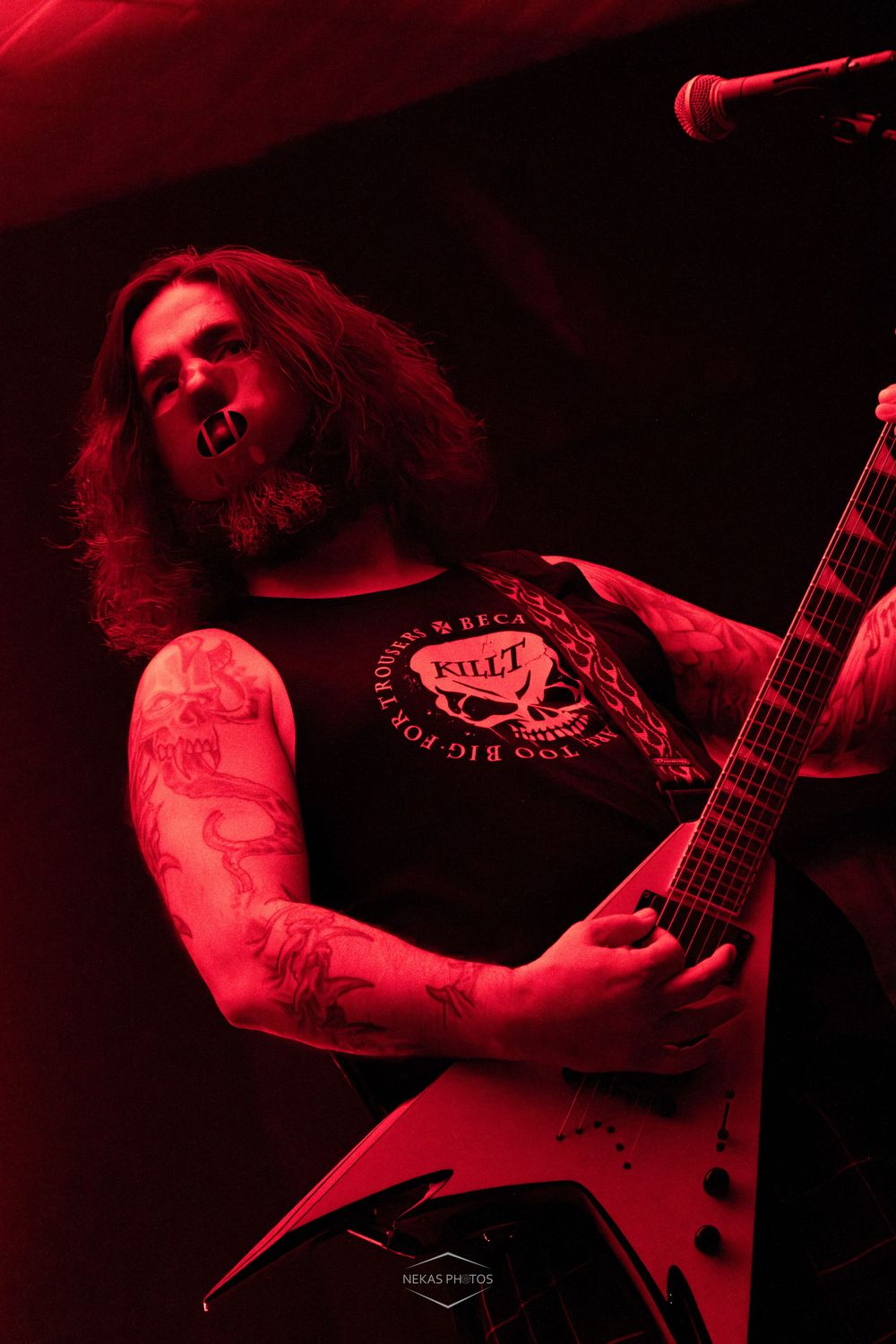 Heavy metal guitarist performs on stage bathed in red light, playing a Flying V guitar while wearing a sleeveless band shirt.