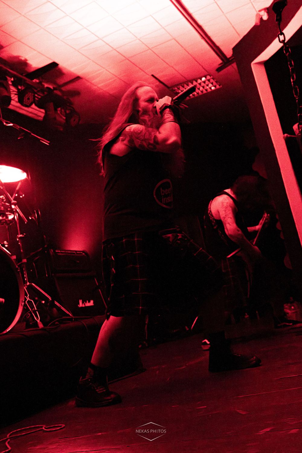 Metal vocalist performing on stage under dramatic red lighting with long hair and band merchandise shirt.