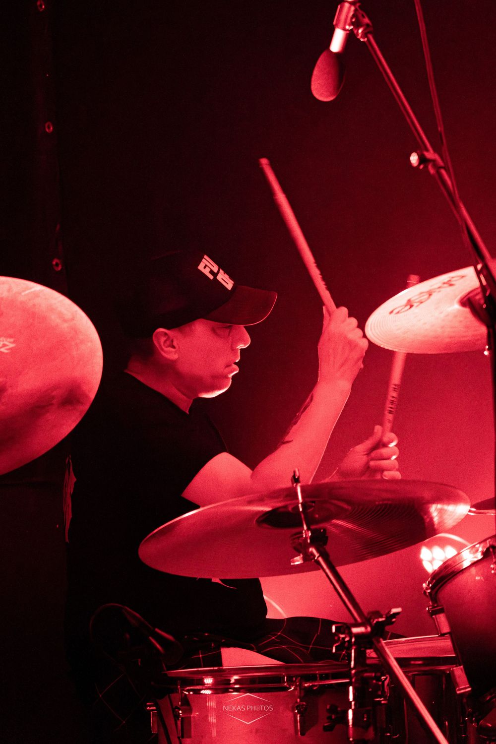 Drummer performing on stage under dramatic red lighting, playing cymbals with drumsticks while wearing a black cap.