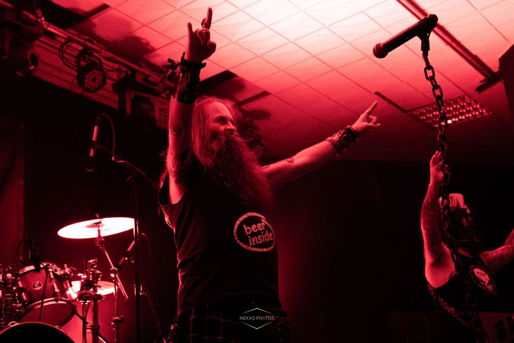 Long-haired metal musician making rock hand gesture on red-lit stage during live concert performance.