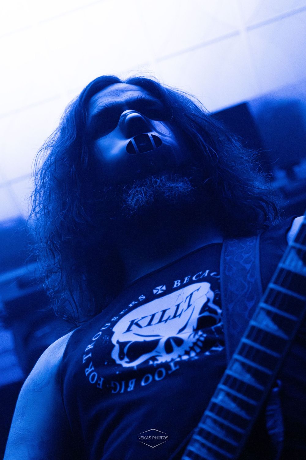 Long-haired metal musician in a skull print tank top performing under dramatic blue stage lighting.