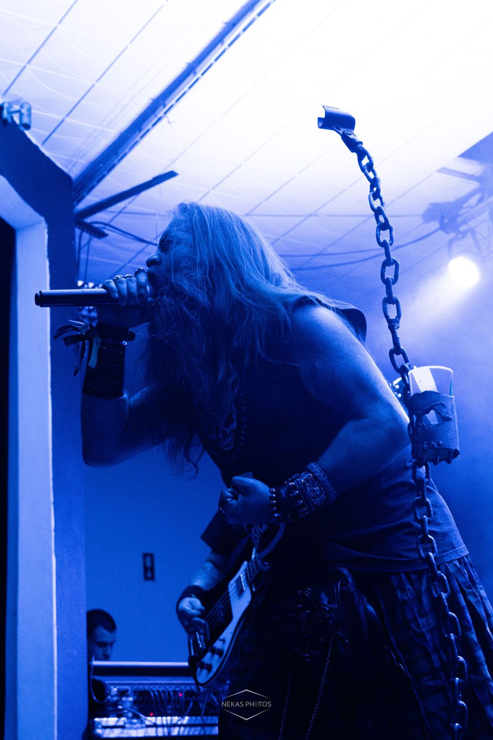 Heavy metal vocalist with long hair performing on stage under blue lighting with chains hanging overhead.