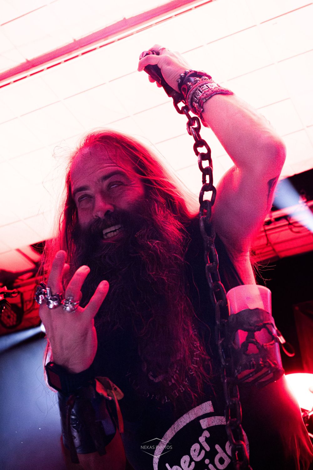 Long-haired bearded man holding a metal chain under red stage lights at a rock concert.