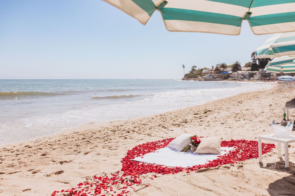 Romantic beach setup with red rose petals in heart shape on white blanket near ocean shore under striped umbrella.