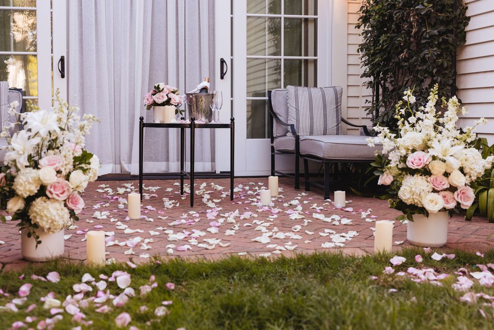 Romantic outdoor patio decorated with white and pink roses, scattered flower petals, and candles for a wedding proposal.
