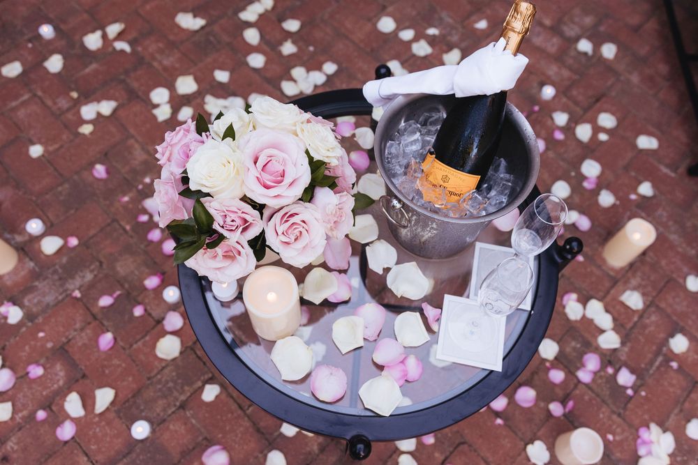 Romantic wedding champagne setup with pink roses bouquet, ice bucket, candles, and rose petals scattered on brick floor.