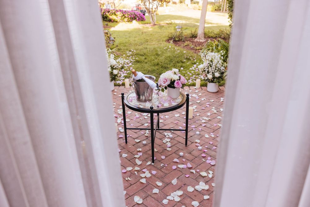 Elegant outdoor wedding setup with a glass table holding flowers, surrounded by rose petals on a brick patio.