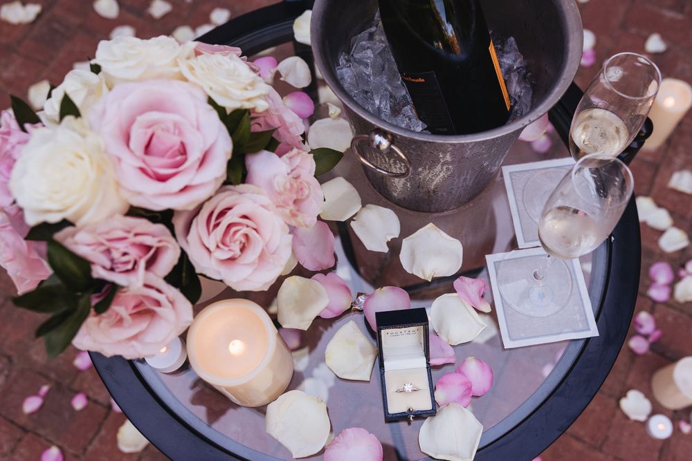 Romantic proposal setup with pink roses, champagne on ice, ring box, candles, and scattered petals on a round tray.