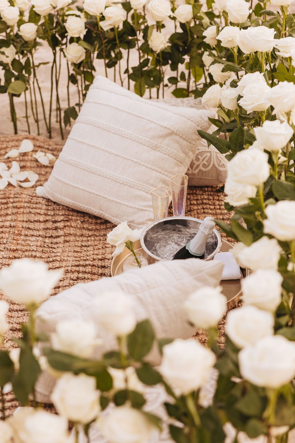 Romantic outdoor setup with white roses, linen cushions, champagne, and a silver bowl on a woven mat.