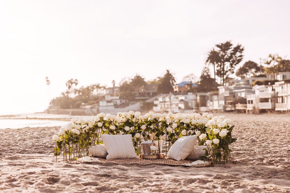 Romantic beach picnic setup with white floral arrangements, pillows, and coastal homes in the background.