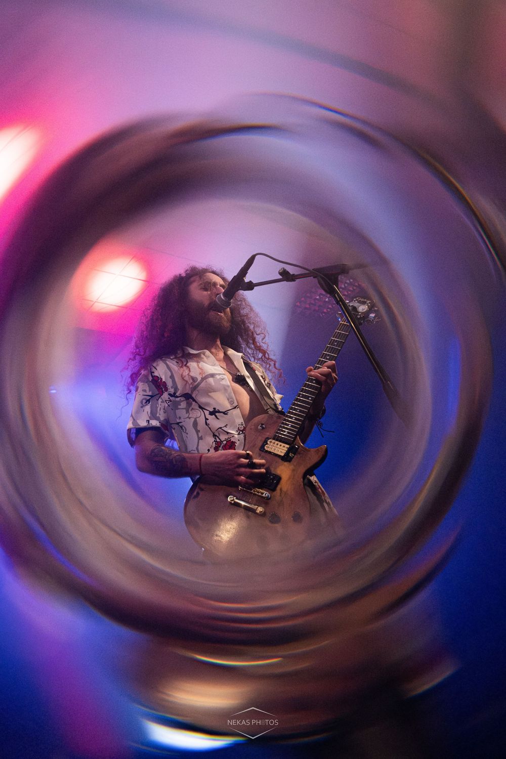 Guitarist with long curly hair performs on stage under colorful lights, captured through a swirling circular lens effect.