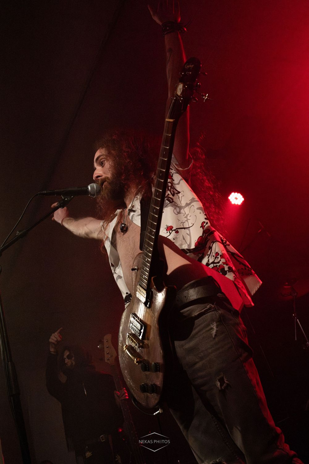 Bearded rock musician singing into mic while holding electric guitar on stage under red lights.