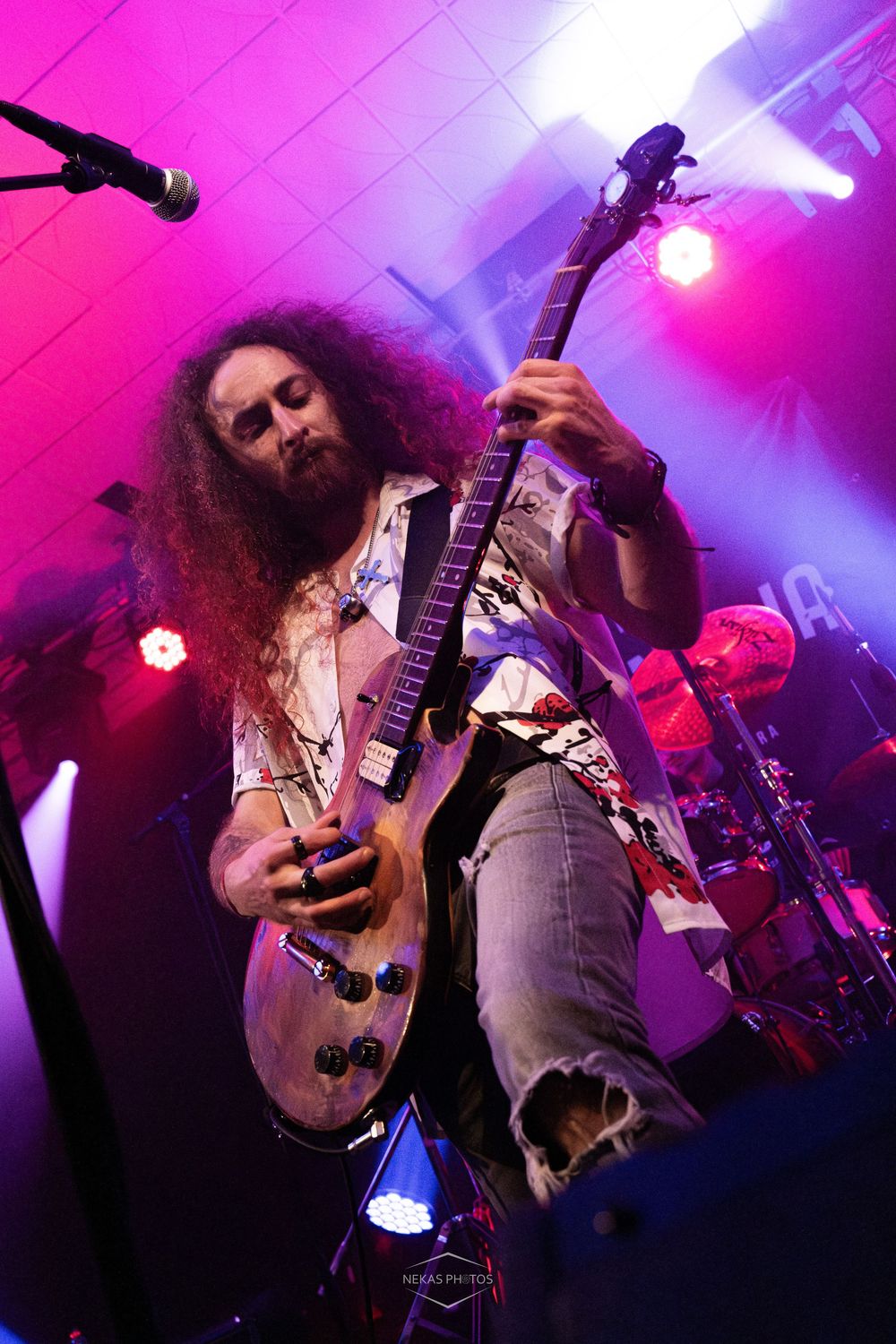 Long-haired bearded guitarist performs on stage under purple lights at a live rock concert.