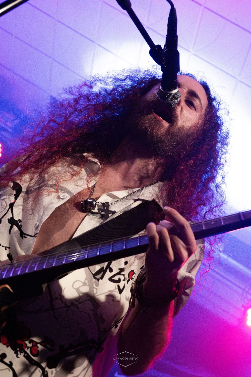 Curly-haired male guitarist singing into microphone on stage under purple lights wearing a white patterned shirt.