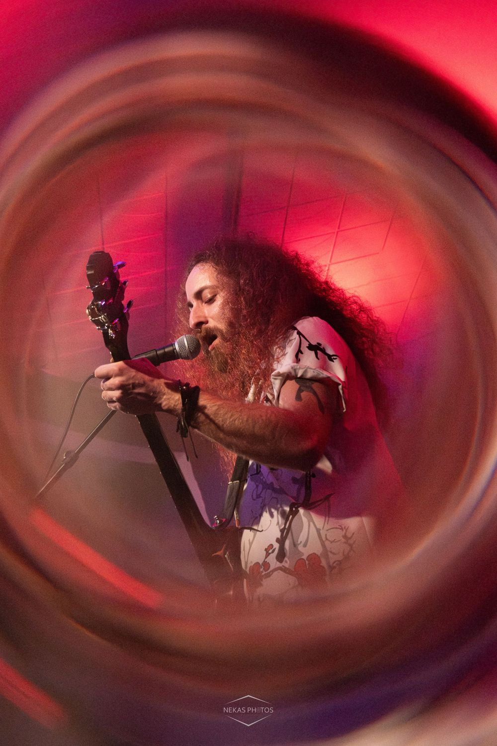 Curly-haired male guitarist singing into microphone on stage, captured through creative circular lens effect with red lighting.