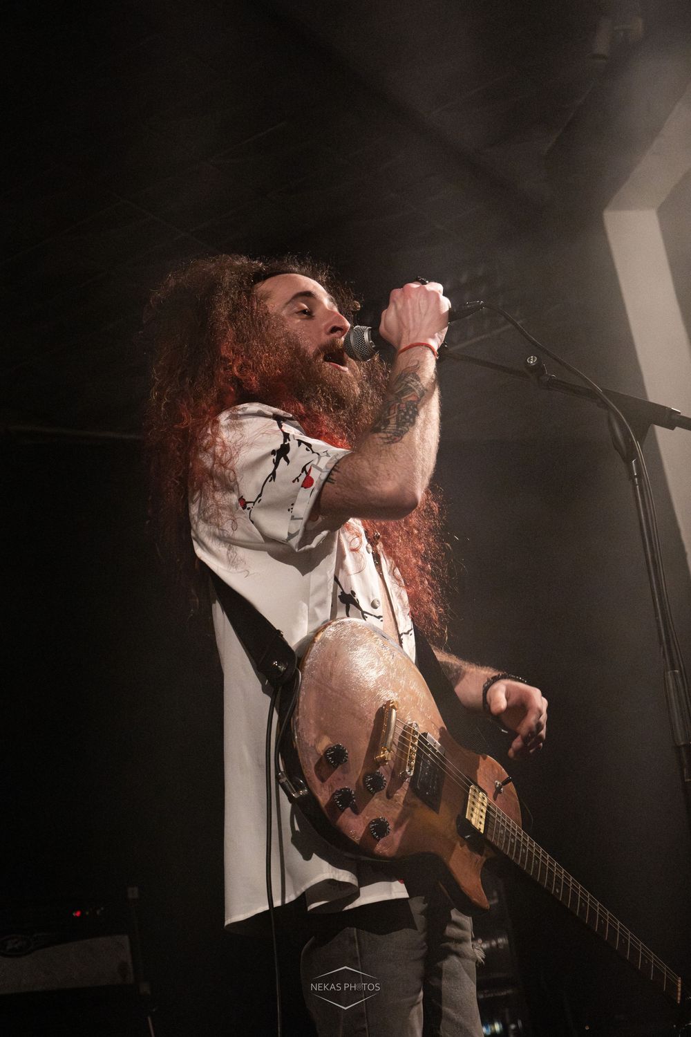 Bearded male rock musician with long red hair singing into microphone while playing electric guitar on dark stage.