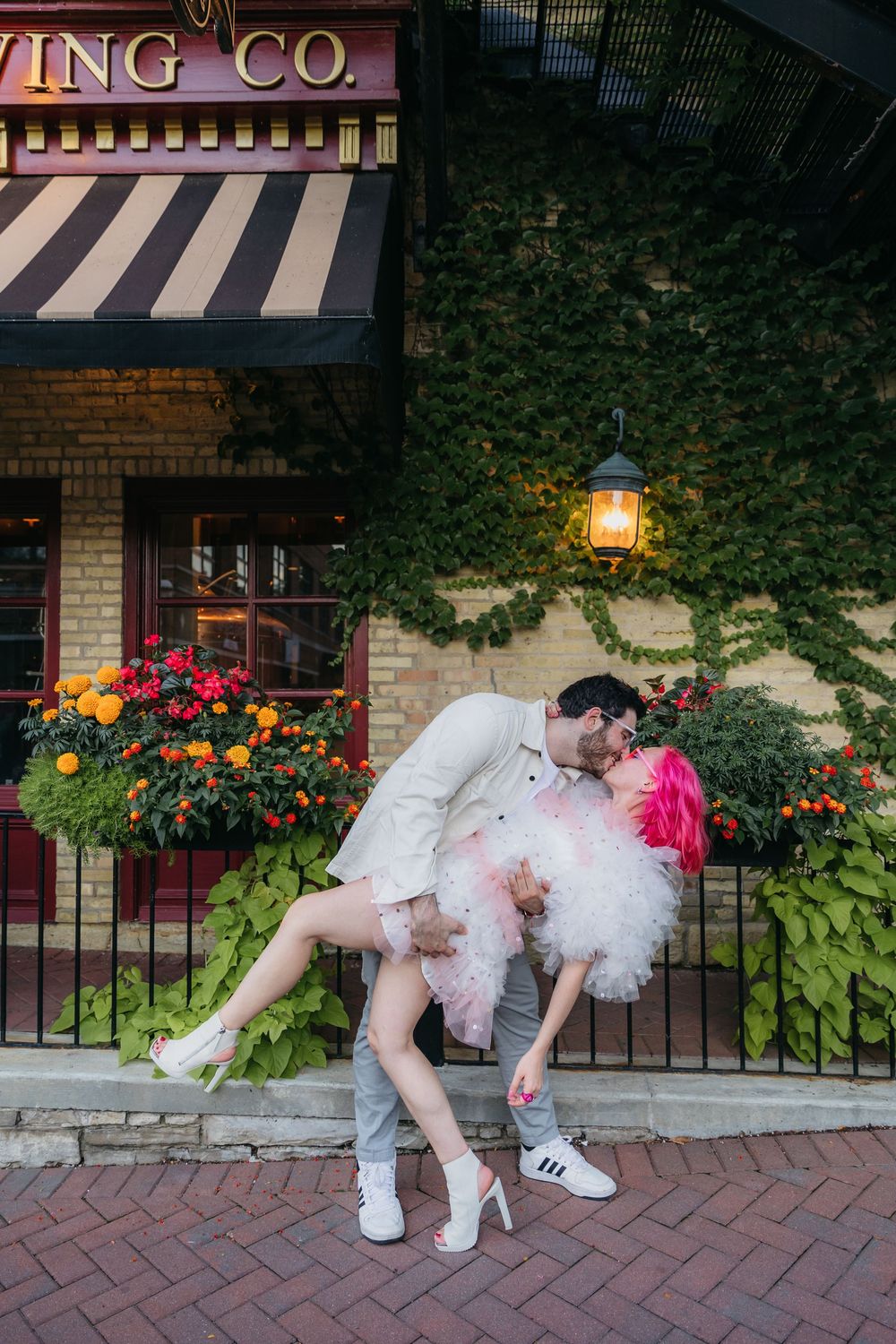 Couple sharing a romantic dip kiss outside a brick building with ivy, colorful flowers, and a lantern in West Dundee, Il