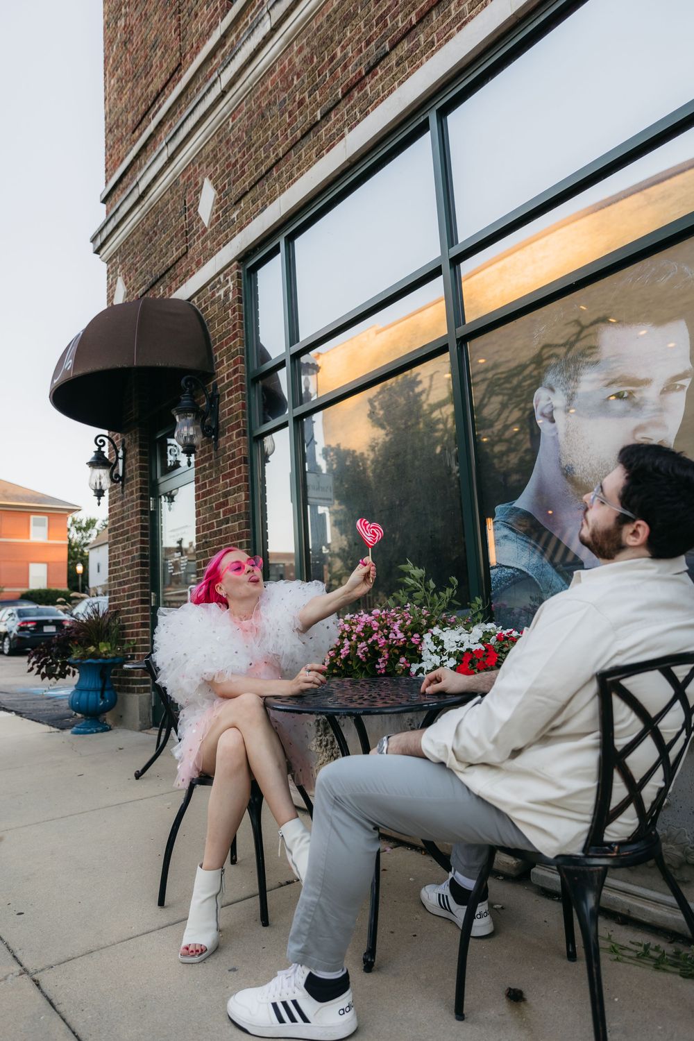 A woman with pink hair holds a heart lollipop while sitting outside a brick building with a man at a cafe in West Dundee during engagement