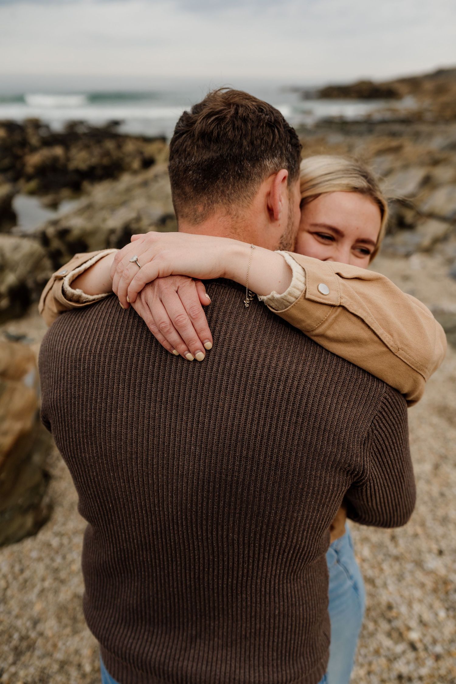 A couple embraces on a rocky beach, the woman smiling as she wraps her arms around a man in a brown sweater.