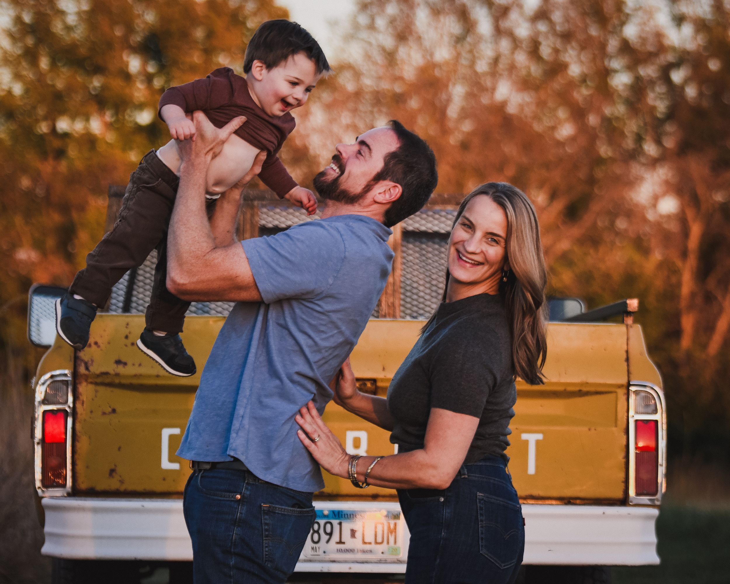 Family photo with parents lifting toddler by vintage yellow truck outdoors.