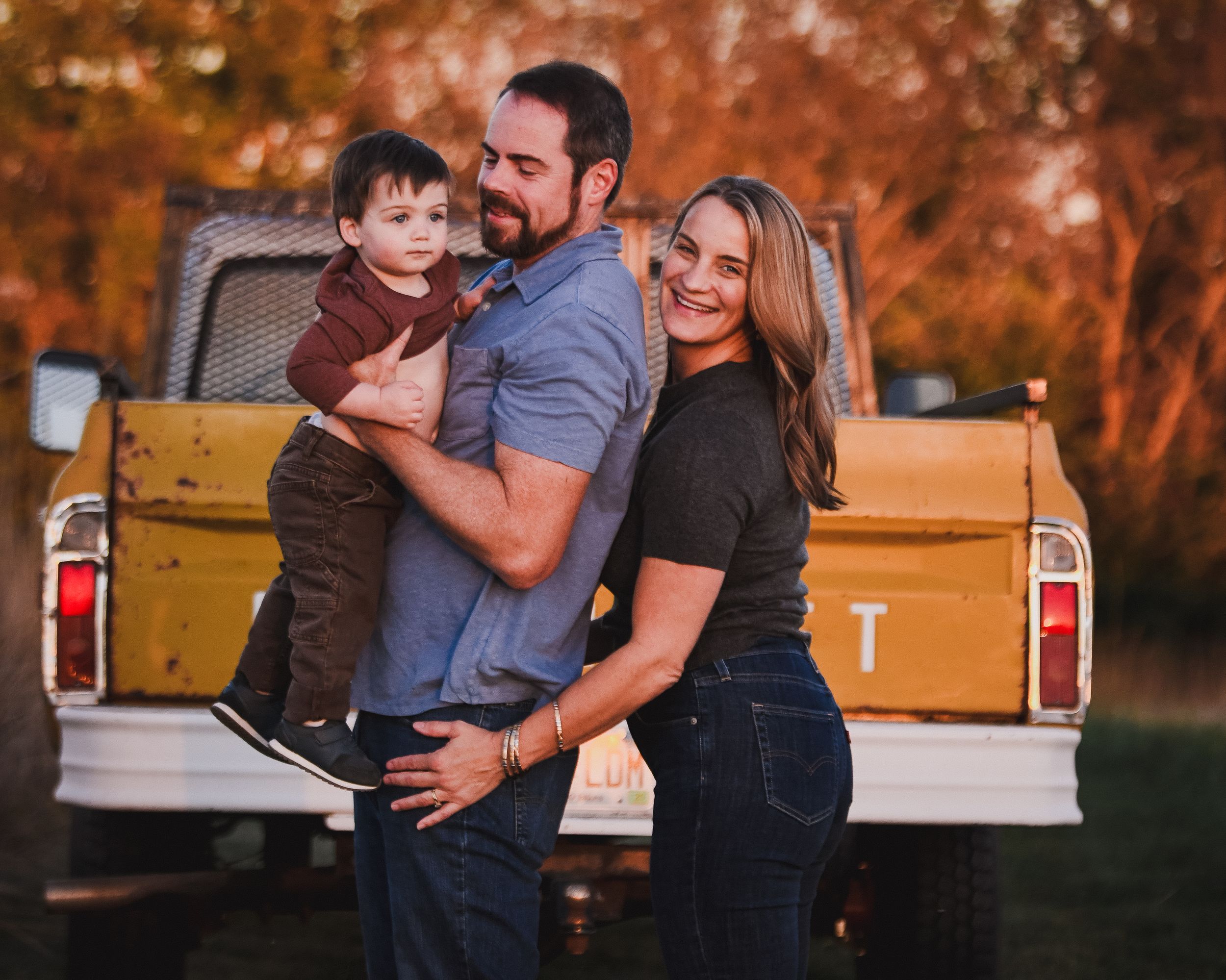 A happy young family with a toddler poses in front of a vintage yellow truck during a golden hour outdoor photo session.