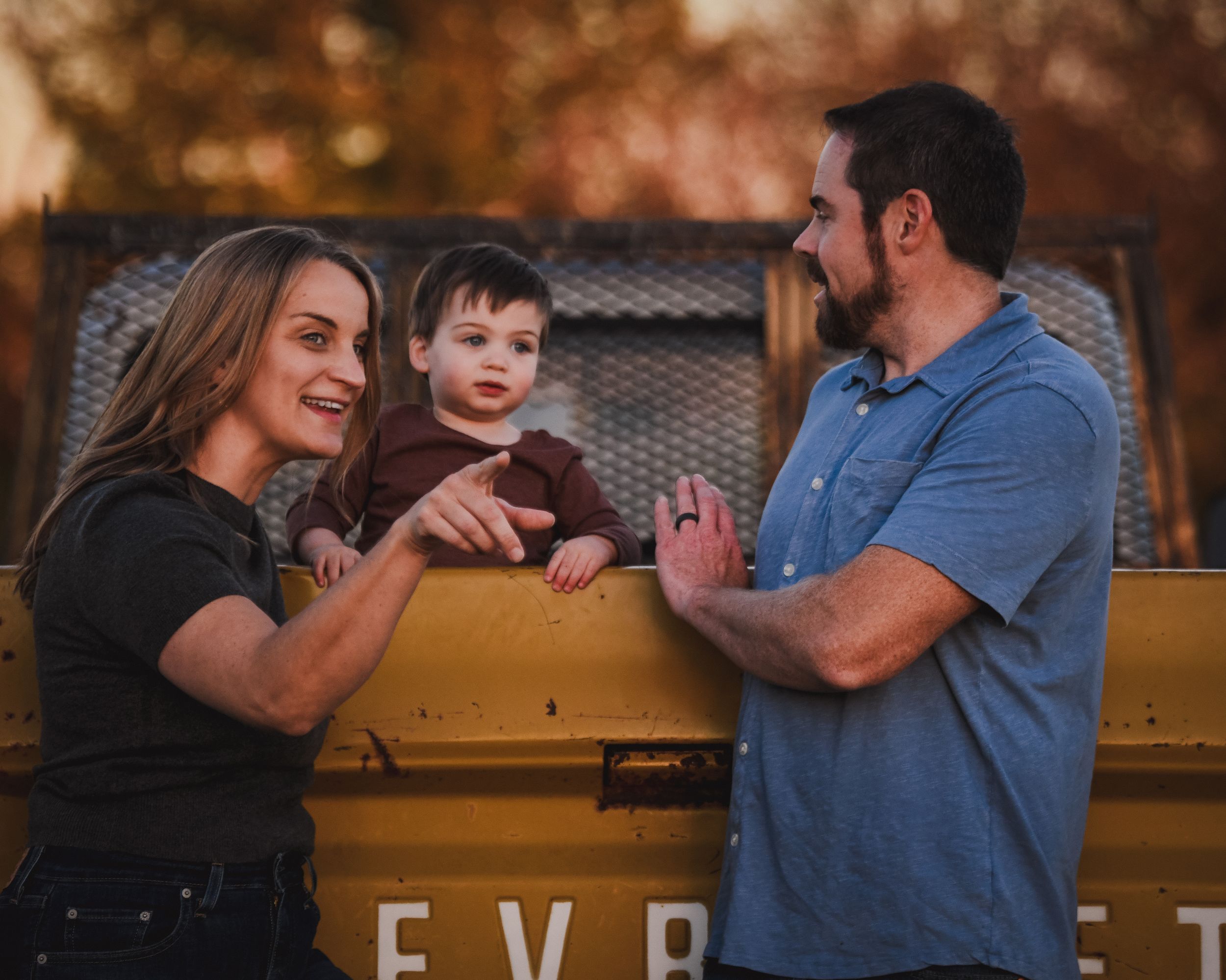 Parents interacting with toddler sitting in yellow vintage truck bed outside.