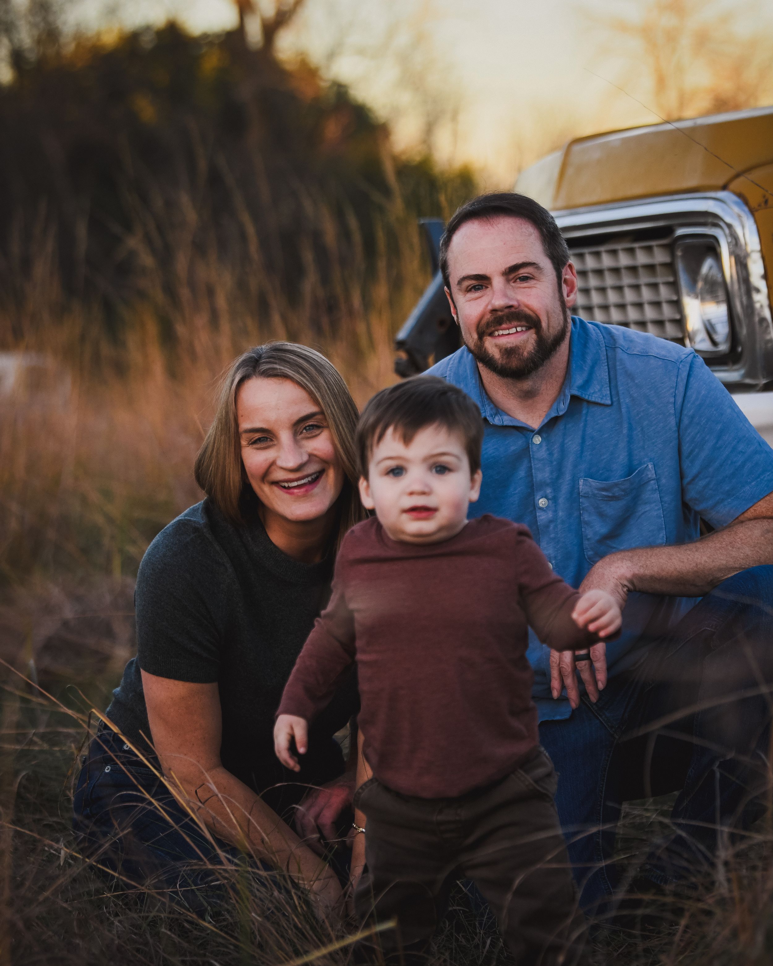Family of three posing outdoors at golden hour near a vintage truck in a field of tall grass.