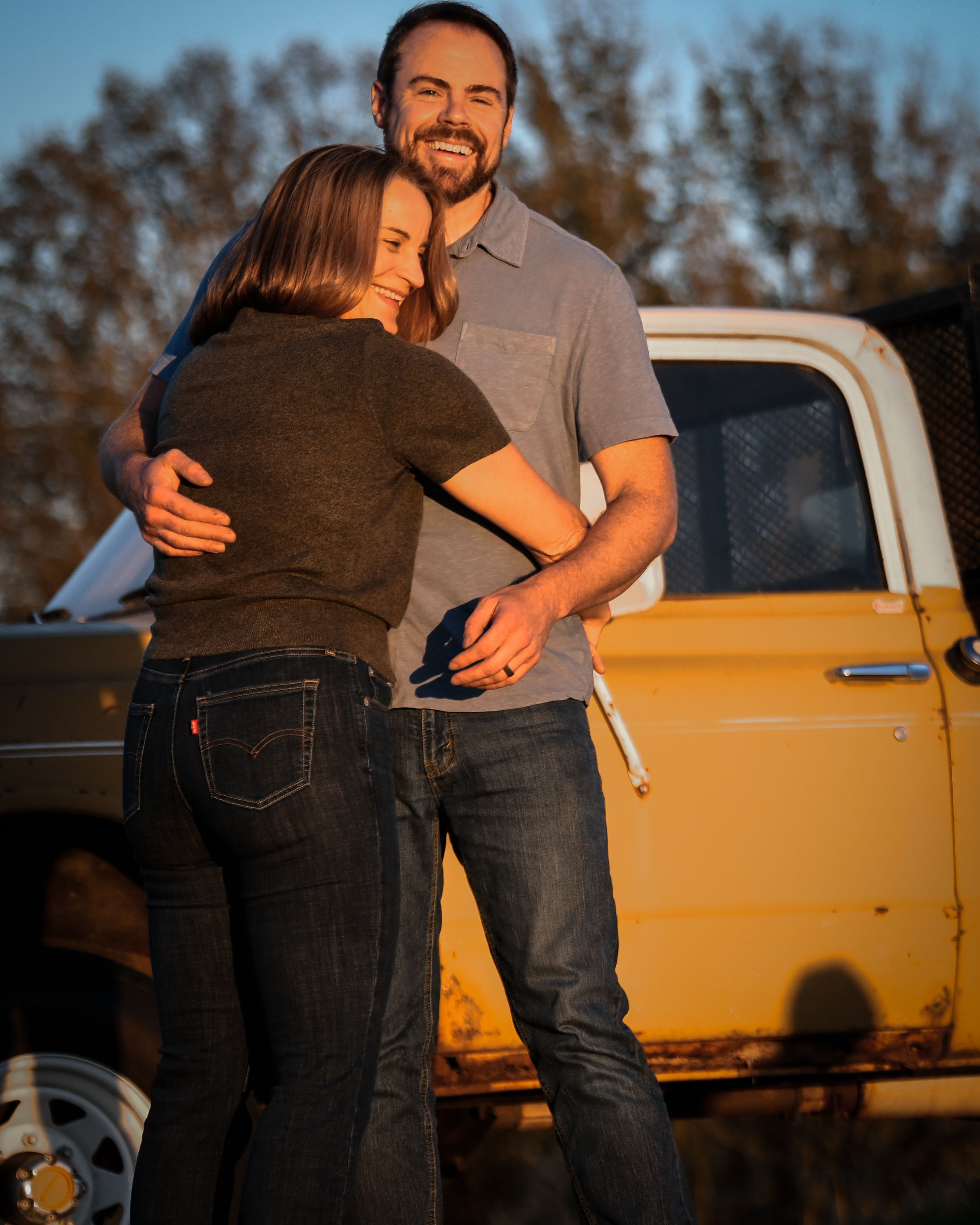 A smiling couple embraces warmly in golden hour light beside a vintage yellow pickup truck outdoors.
