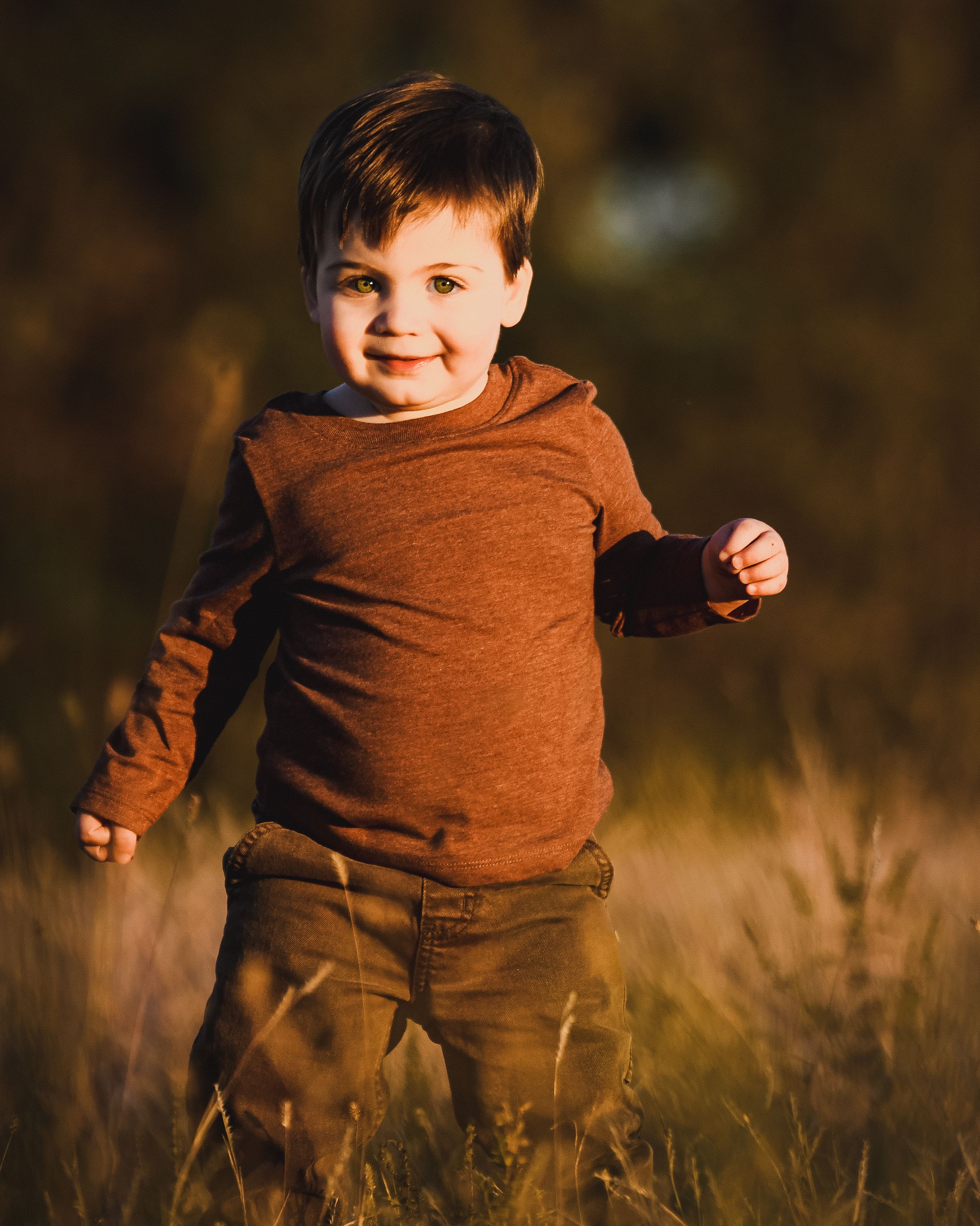 A cheerful toddler boy in a brown hoodie and khaki pants runs through a sunlit golden grass field.