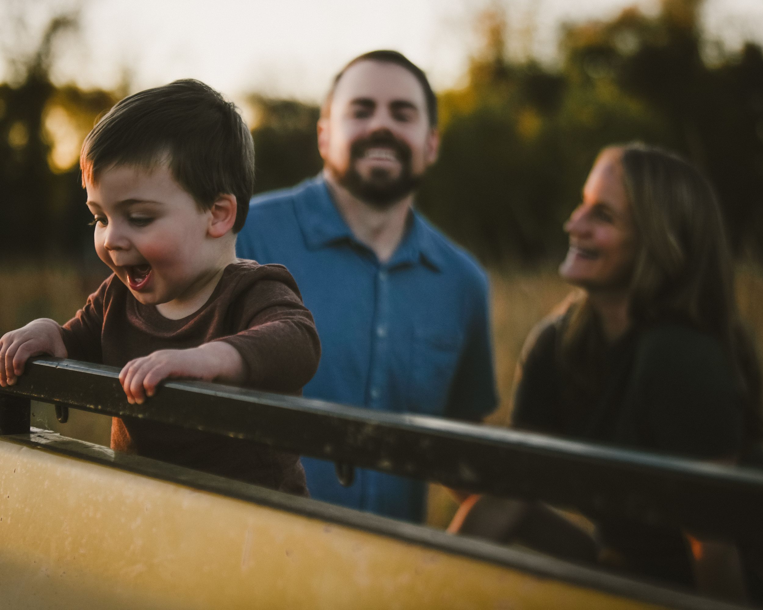 Toddler laughing at edge of yellow truck bed with parents smiling behind him.