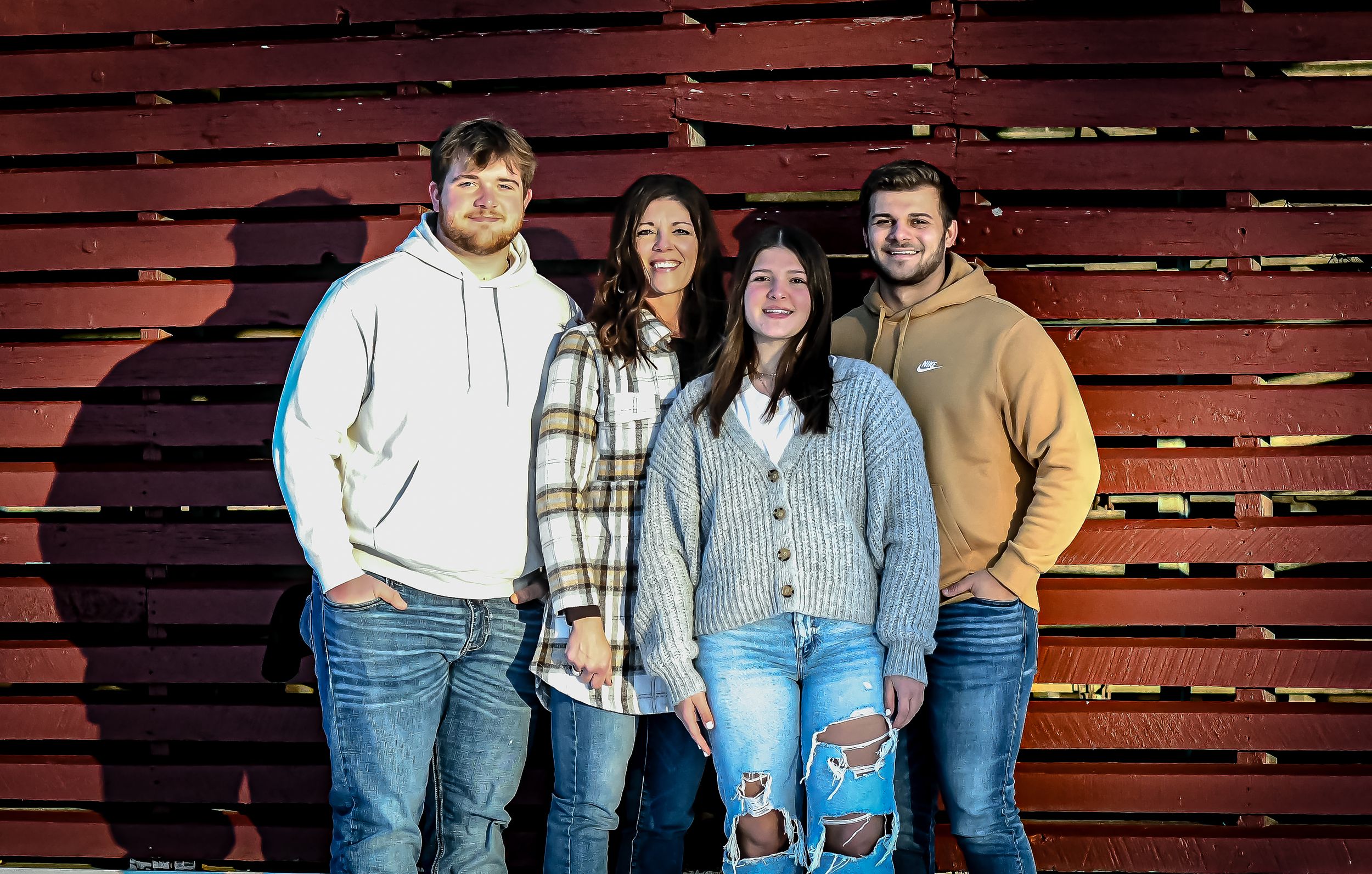 Four smiling friends pose together in front of a rustic red wooden wall wearing casual jeans and hoodies.