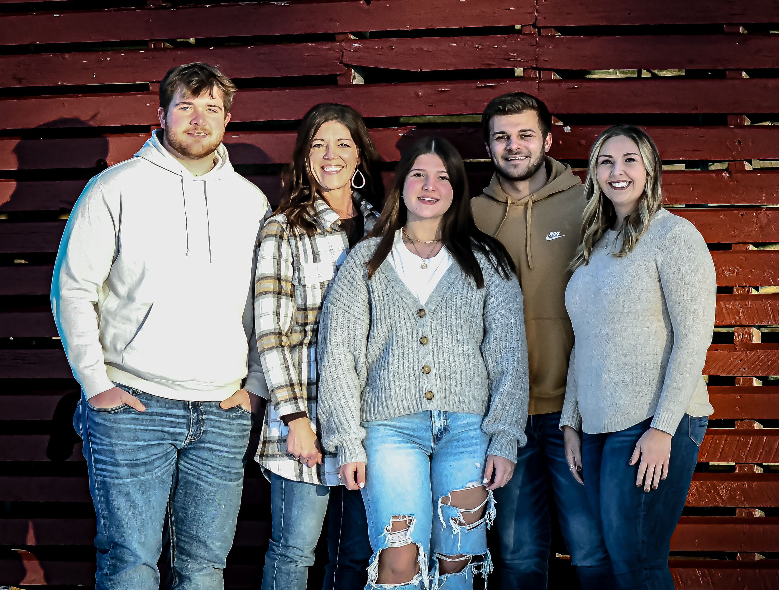 Five young adults smiling together in front of a rustic red barn wood wall in casual winter clothing.